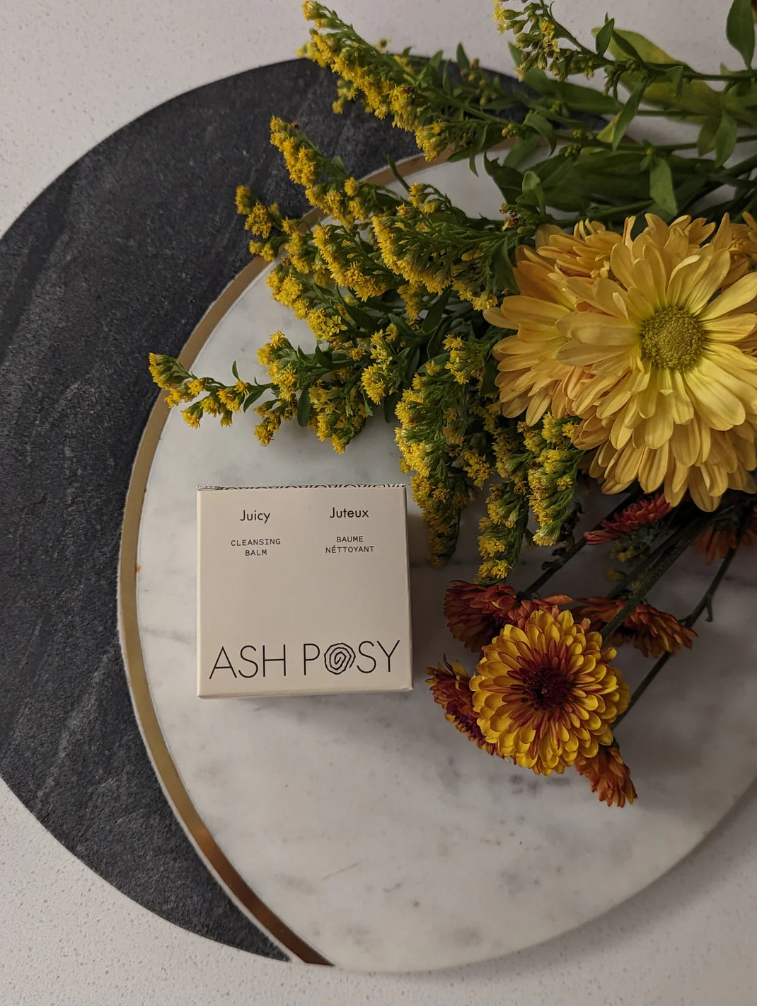 a styled photo of an Ash Posy skincare product surrounded by yellow flowers