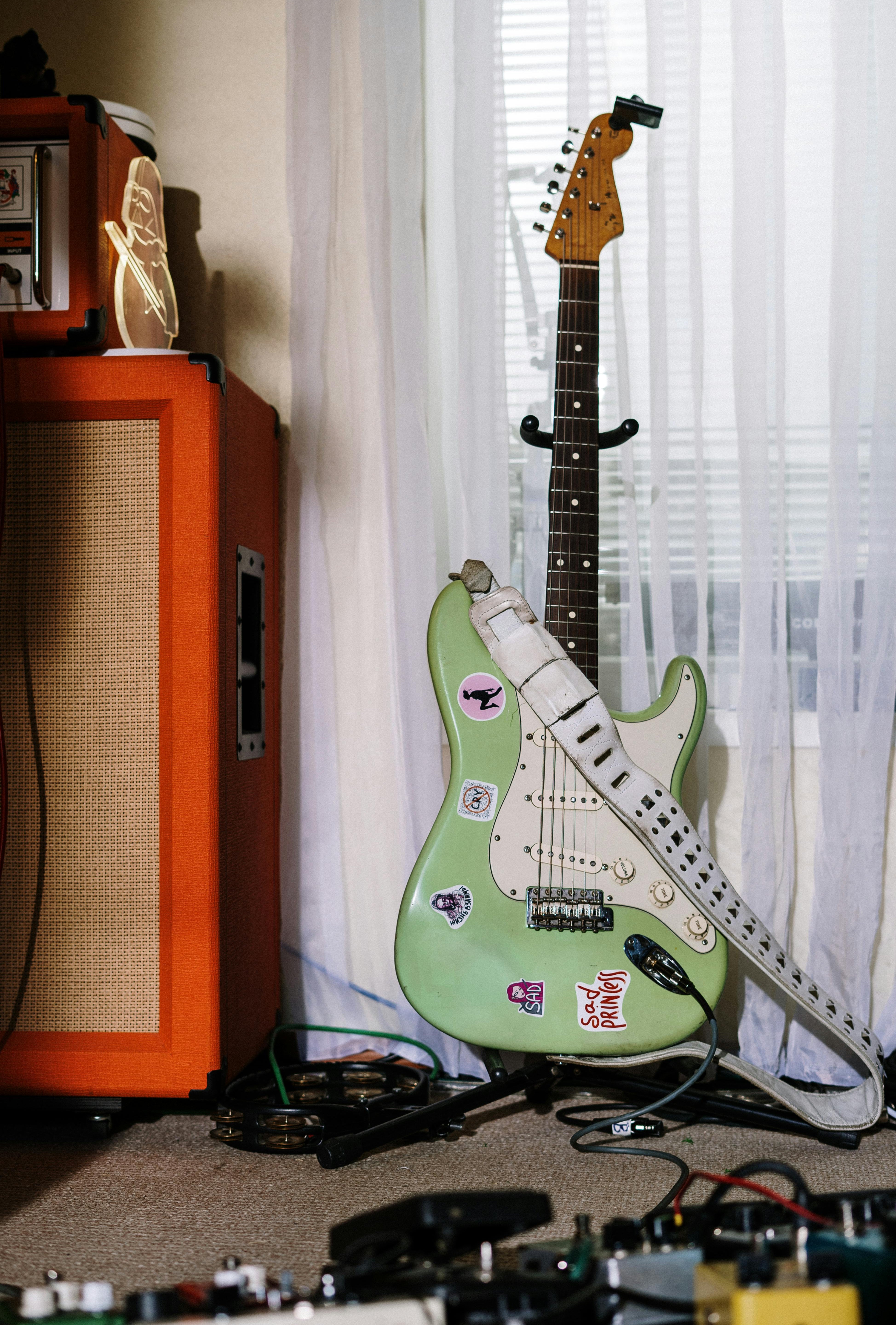 Green electric guitar with stickers next to an orange amplifier in a room with white curtains.